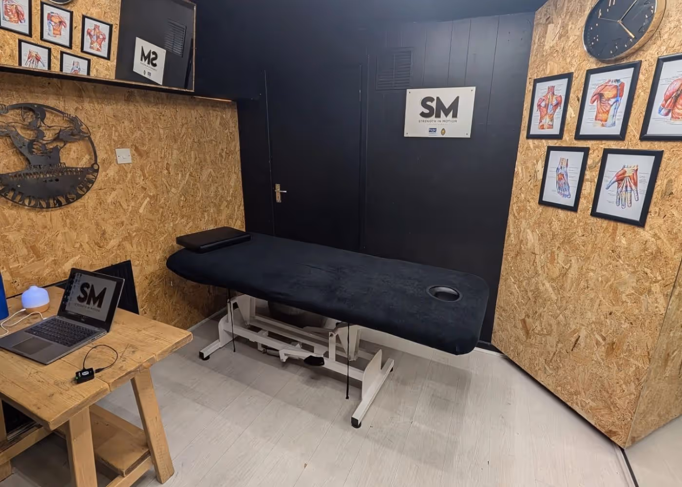 Massage therapy room with a black massage table, wooden desk with a laptop, anatomical muscle charts on walls, and a clock.